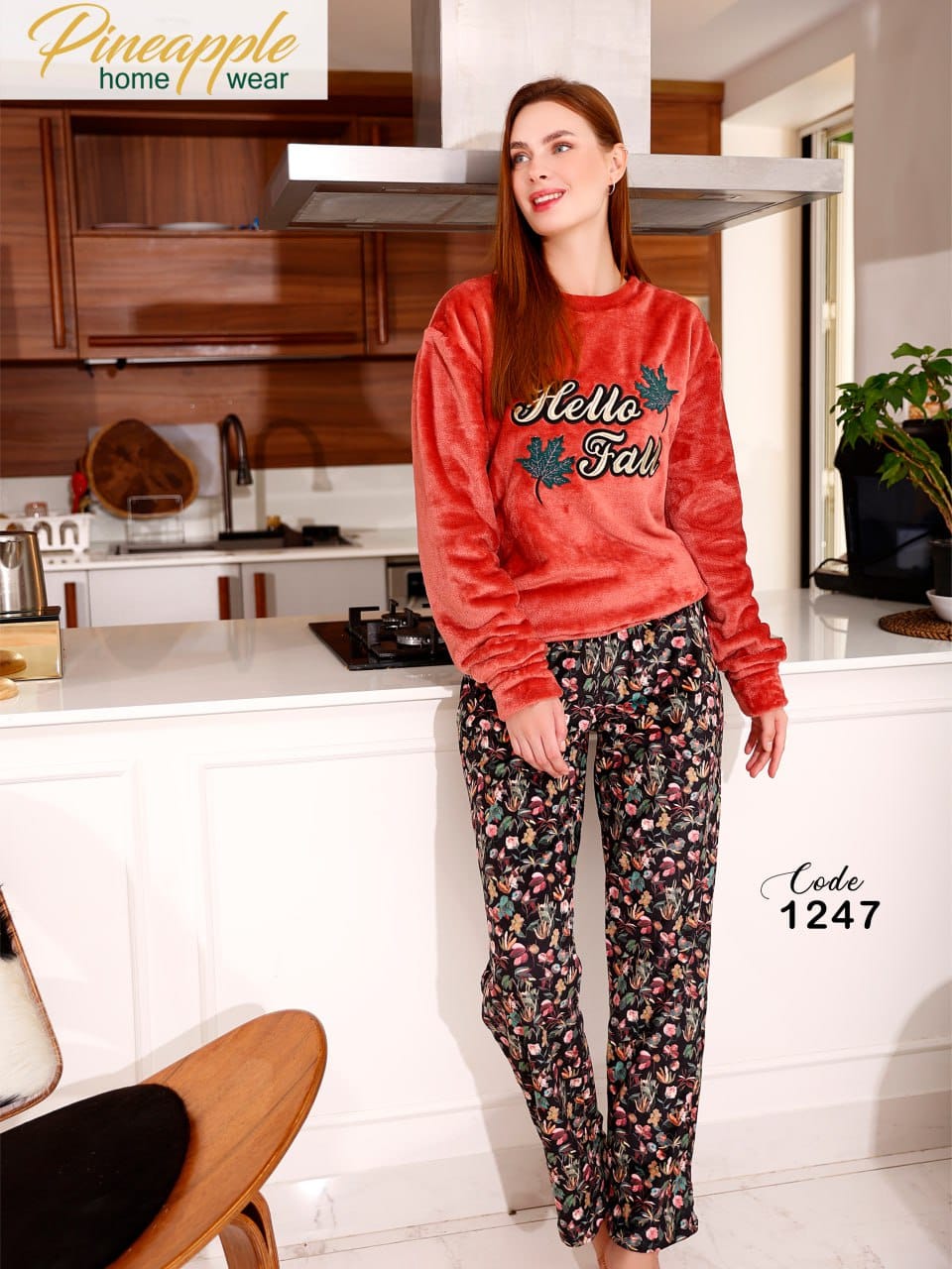 Woman wearing a red 'Hello Fall' sweater and floral pants in a kitchen setting, with Pineapple Home Wear branding.