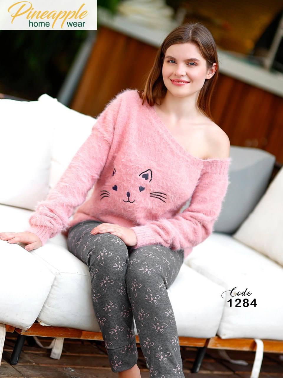 Woman wearing a pink sweater with a cat design and gray pants, sitting on a white couch.