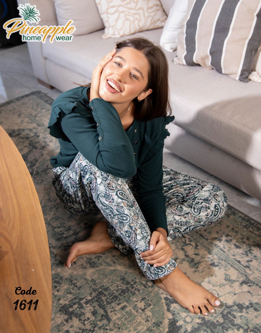 Woman sitting on a rug in a living room wearing a green top and patterned pants, with 'Pineapple Home Wear' branding.