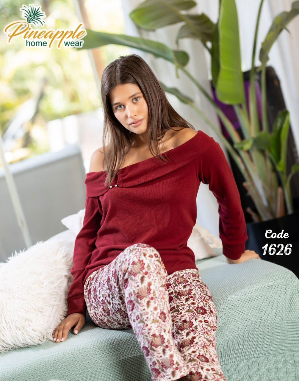Woman wearing a red off-shoulder top and patterned pants sitting on a couch with plants in the background.
