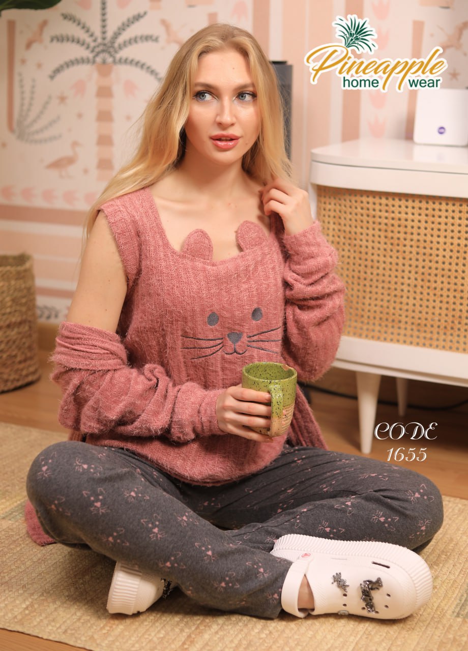 Woman wearing a pink cat-themed sweater and gray pants, sitting on the floor with a cup, in a room with wicker furniture and patterned walls.