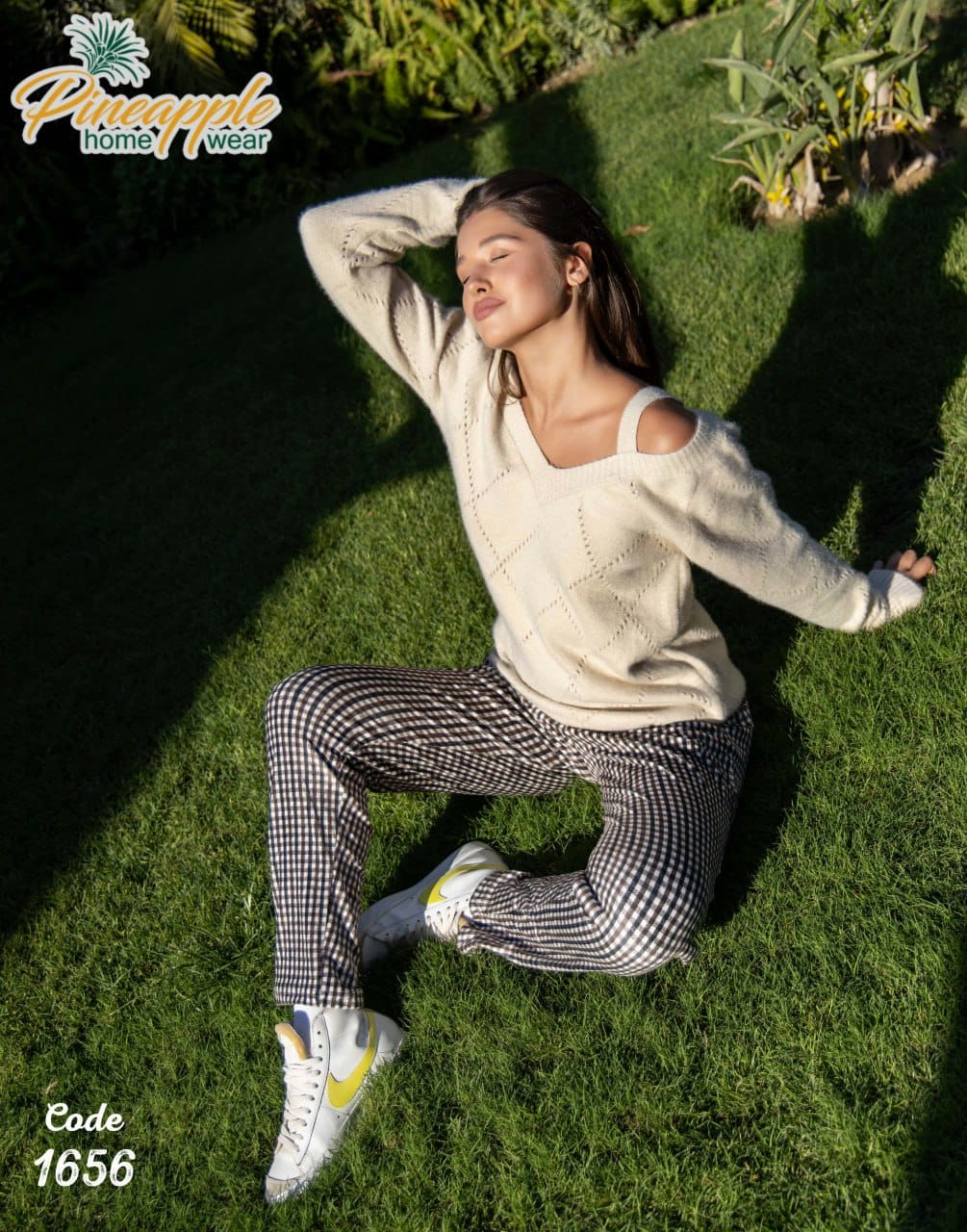 Woman lying on grass wearing a white sweater and checkered pants with 'Pineapple Home Wear' branding.