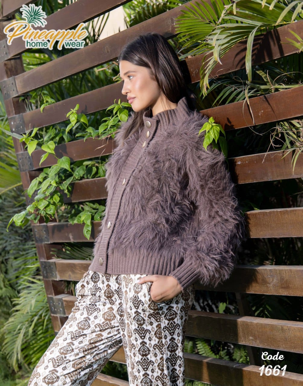 Woman wearing a textured brown cardigan and patterned pants leaning against a wooden wall with plants.