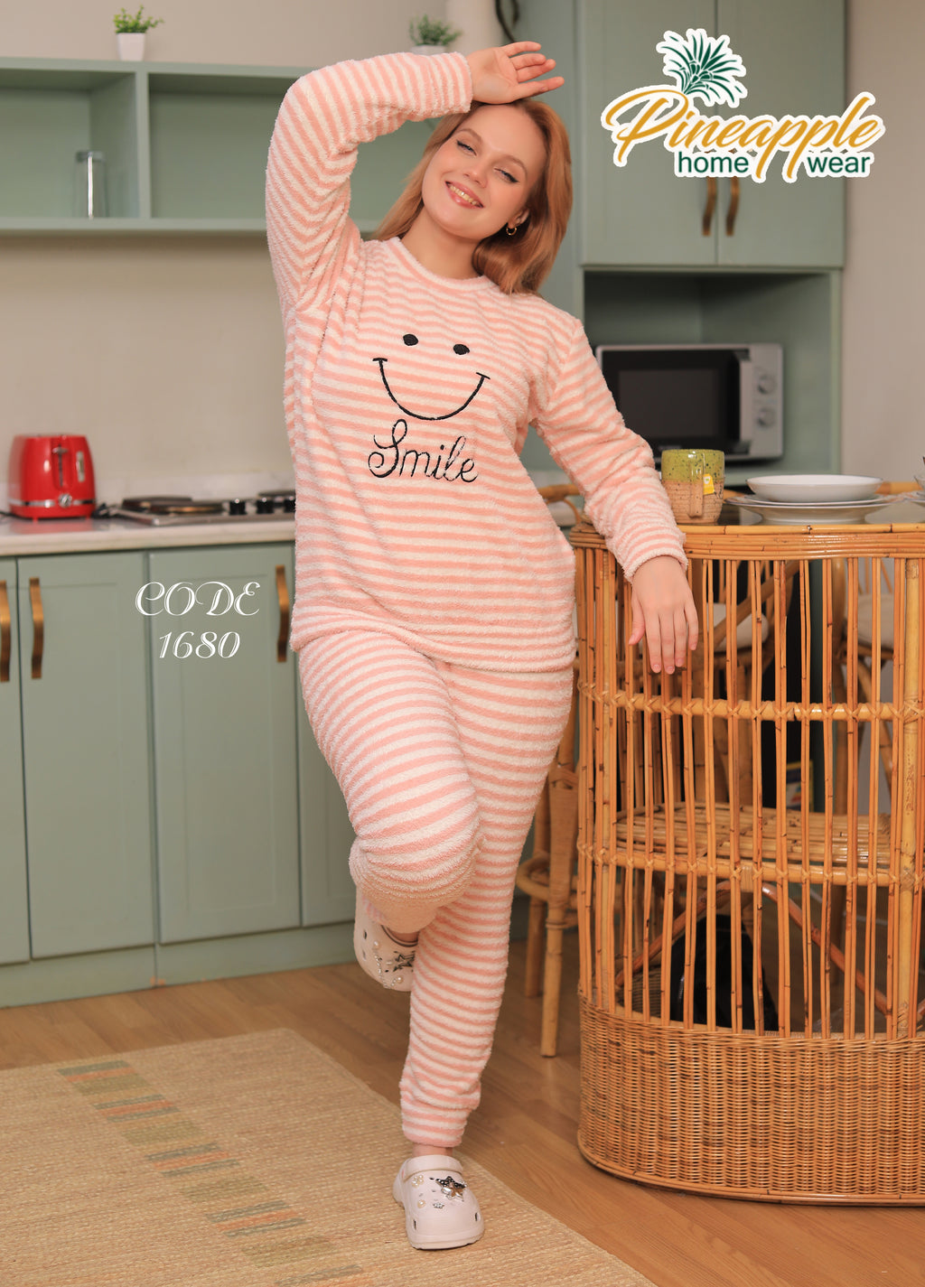 Woman wearing a pink smiley face pajama set in a kitchen, with 'Pineapple Home Wear' branding.