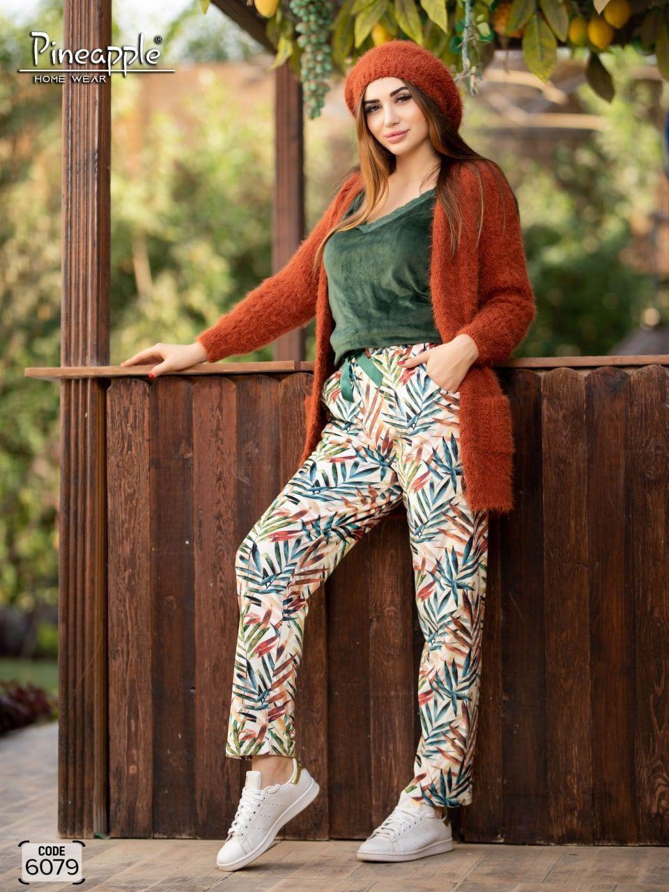 Woman wearing a rust-colored cardigan, green top, and colorful pants leaning against a wooden fence.