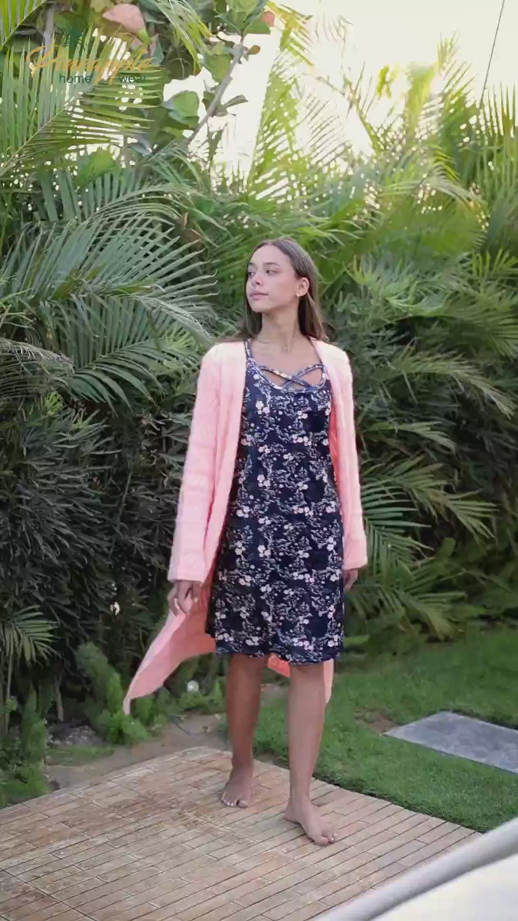 Woman wearing a navy floral dress with a pink cardigan against a green leafy background, branded 'Pineapple Home Wear'.
